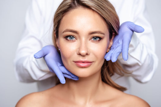 Close up portrait of young blonde woman with cosmetologyst hands in a gloves. Preparation for operation or procedure. Perfect skin, spa and care